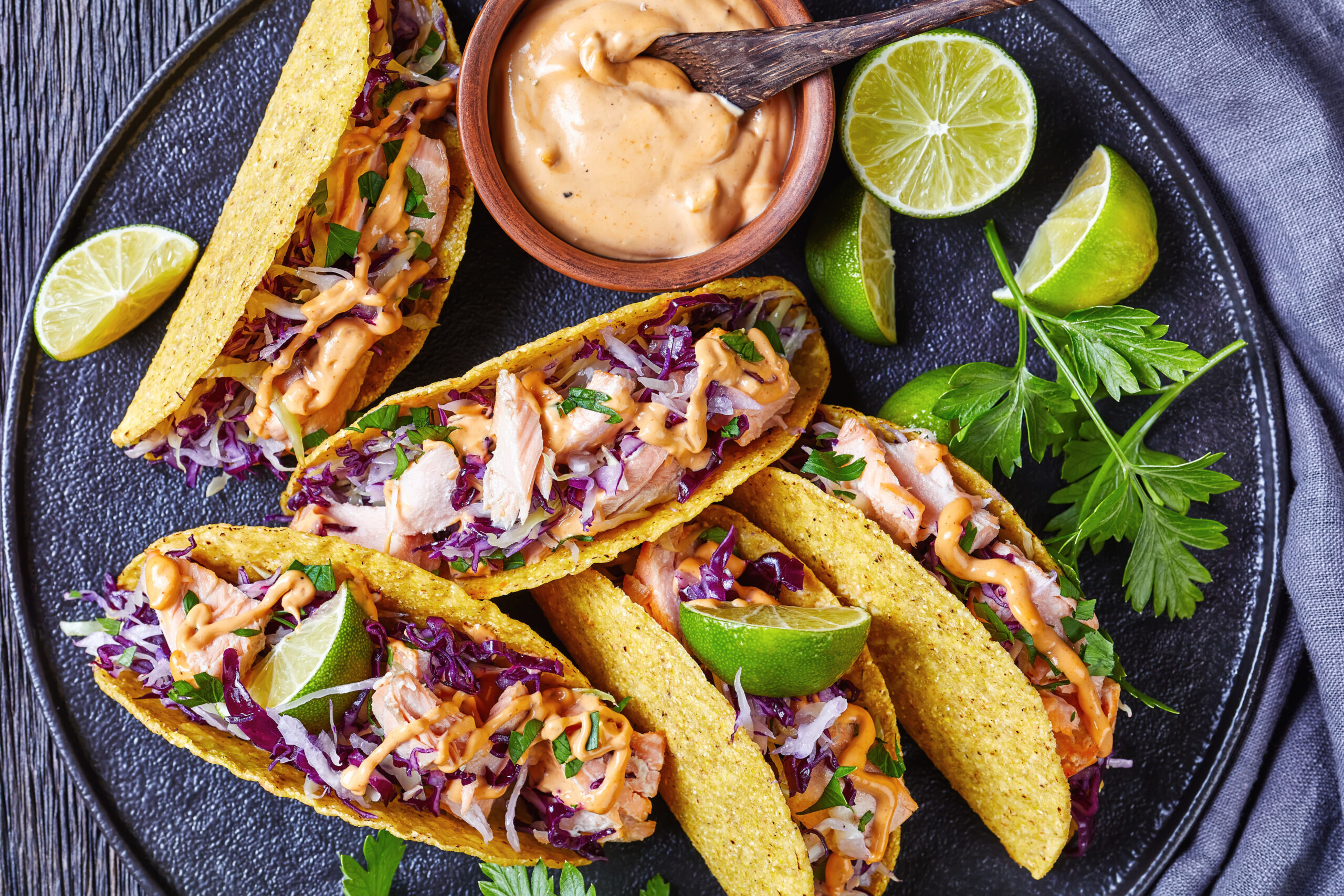 salmon tacos with red cabbage salad with spicy yogurt sauce sprinkled with finely chopped parsley served on a black plate on a dark wooden table, horizontal view from above, flat lay, close-up